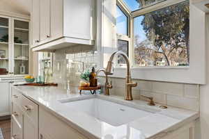 Kitchen view of white cabinetry, light stone countertops, tasteful backsplash, and glass insert cabinets