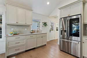 Kitchen with vaulted ceiling, stainless steel fridge with ice dispenser, light wood finished floors, decorative backsplash, and recessed lighting
