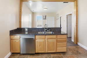 Kitchen with light wood finish cabinetry, stainless steel dishwasher, a peninsula, dark stone finish flooring, and dark stone counters