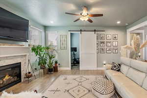 Living area with a barn door, ceiling fan, a brick fireplace, light wood-type flooring, and recessed lighting