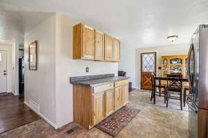 Kitchen with stone finish flooring, freestanding refrigerator, dark countertops, light wood finish cabinetry, and a textured ceiling