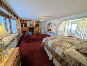 Bedroom featuring a glass covered fireplace, dark carpet, and a ceiling fan