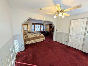 Bedroom with a wainscoted wall, a decorative wall, dark colored carpet, a ceiling fan, and arched walkways