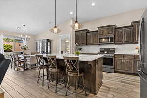 Kitchen with dark wood finish cabinetry, stainless steel appliances, a center island with sink, vaulted ceiling, and light stone countertops