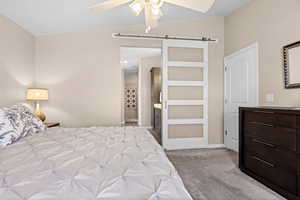 Bedroom with a barn door, light colored carpet, lofted ceiling, ensuite bathroom, and a ceiling fan