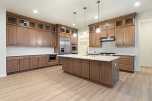 Kitchen featuring wood finish cabinetry, stainless steel appliances, a center island with sink, decorative light fixtures, and light stone counters