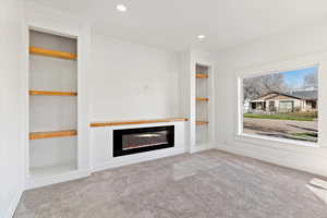 Unfurnished living room featuring built in shelves, light carpet, a glass covered fireplace, and recessed lighting