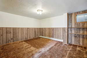 Empty room featuring wood walls, a textured ceiling, wainscoting, a baseboard radiator, and dark carpet