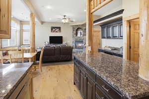 Kitchen featuring light wood-type flooring, open floor plan, a stone fireplace, a ceiling fan, and dark stone countertops