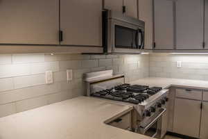 Kitchen with gray cabinetry, stainless steel appliances, backsplash, and light stone countertops