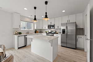 Kitchen featuring stainless steel appliances, white cabinetry, light LVP flooring, and a center island. Quartz countertops, pendant lighting, and a subway tile backsplash complete the space.