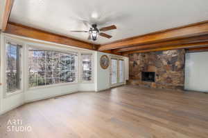 Unfurnished living room with a ceiling fan, light wood finished floors, beamed ceiling, a stone fireplace, and a textured ceiling