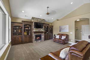 Living area with arched walkways, ceiling fan, carpet floors, vaulted ceiling, and a stone fireplace