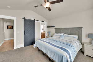 Bedroom with a barn door, vaulted ceiling, light carpet, a ceiling fan, and ensuite bathroom