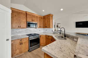 Kitchen featuring stainless steel appliances, lofted ceiling, wood finish cabinetry, light stone countertops, and arched walkways