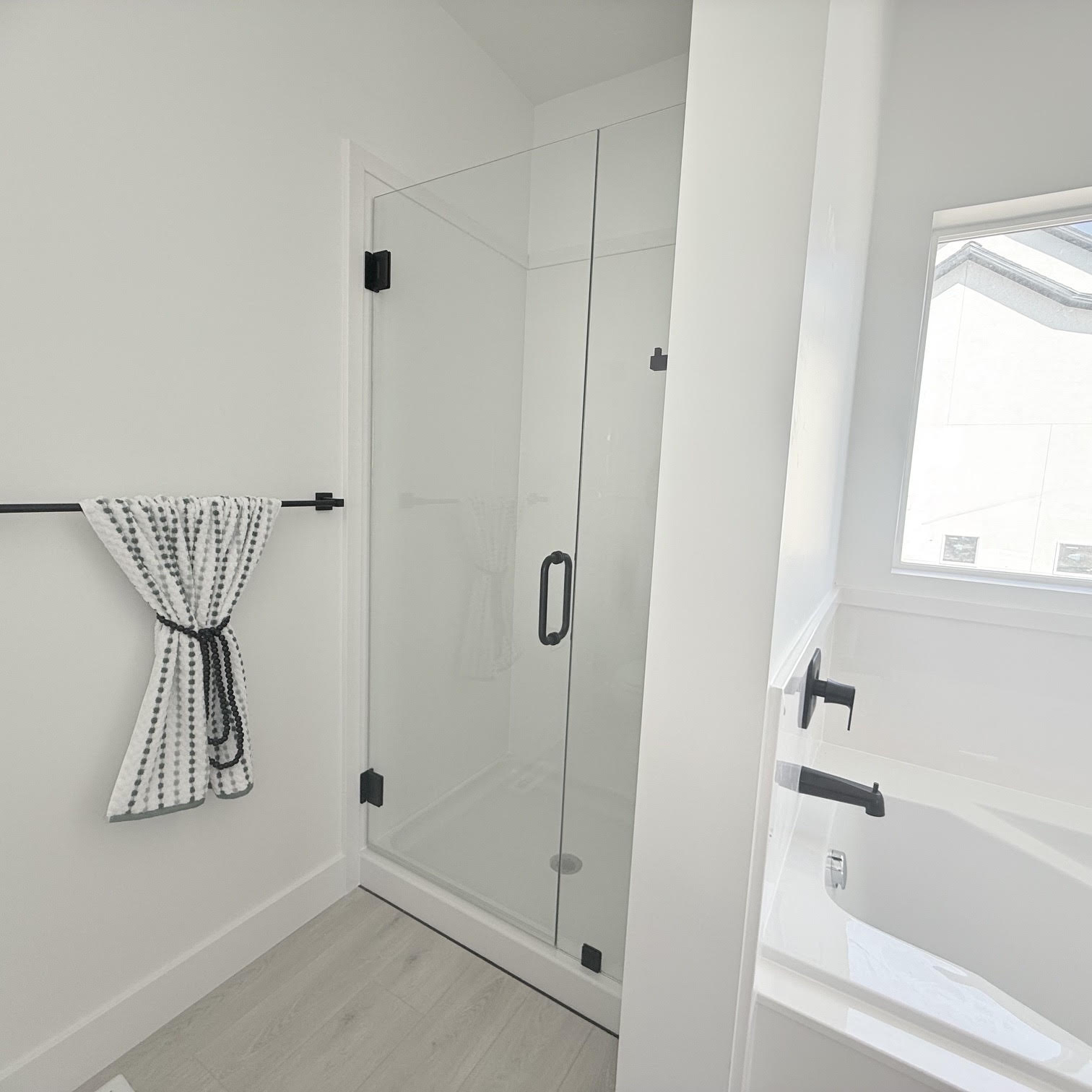 Modern bathroom featuring a frameless glass shower enclosure with matte black hardware, a pristine white tub with matte black fixtures, light-toned wood-finish flooring, and a white baseboard trim