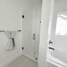 Modern bathroom featuring a frameless glass shower enclosure with matte black hardware, a pristine white tub with matte black fixtures, light-toned wood-finish flooring, and a white baseboard trim