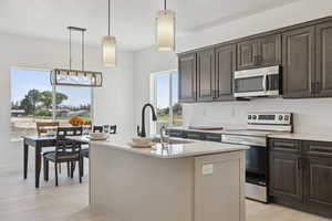 Kitchen with stainless steel appliances, light wood-type flooring, decorative light fixtures, light stone counters, and a kitchen island with sink