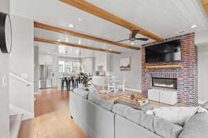 Living room featuring light wood-type flooring, a ceiling fan, recessed lighting, beamed ceiling, and a brick fireplace