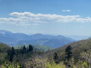 Expansive mountain landscape featuring distant peaks, verdant hillsides, and a clear blue sky with scattered clouds