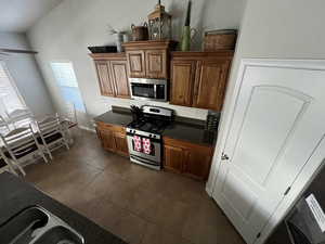 Kitchen with stainless steel appliances, wood finish cabinets, dark tile patterned flooring, vaulted ceiling, and dark stone counters