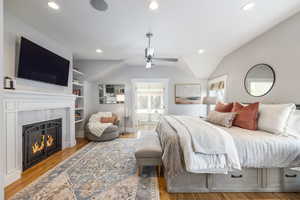 Bedroom featuring light wood-type flooring, a ceiling fan, ensuite bathroom, a lit fireplace, and vaulted ceiling