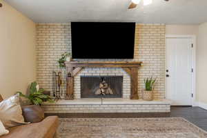 Living room with a brick fireplace, dark wood finished floors, ceiling fan, and a textured ceiling