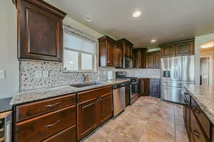 Kitchen featuring stainless steel appliances, dark wood finish cabinets, decorative backsplash, light stone counters, and recessed lighting