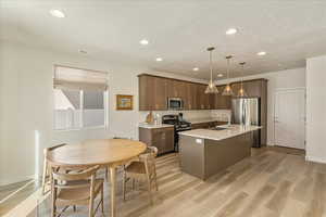 Kitchen featuring stainless steel appliances, a center island with sink, light wood-style floors, pendant lighting, and light stone countertops