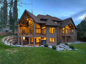 Back of house at dusk with a lawn, a patio area, stucco siding, a balcony, and a shingled roof