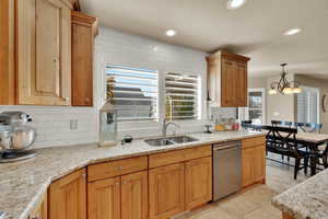 Kitchen featuring backsplash, light stone countertops, stainless steel dishwasher, and suspended lighting