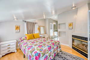 Bedroom featuring vaulted ceiling, a glass covered fireplace, and light wood finished floors