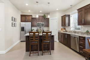Kitchen featuring stainless steel appliances, dark wood finish cabinetry, light stone counters, and an island with sink