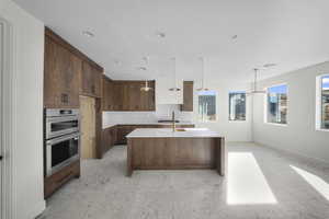 Kitchen featuring an island with sink, light stone countertops, double oven, decorative light fixtures, and light floors
