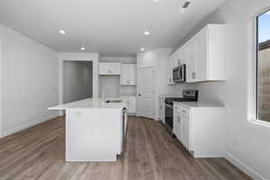 Kitchen featuring white cabinetry, stainless steel appliances, an island with sink, light wood-style flooring, and light stone counters