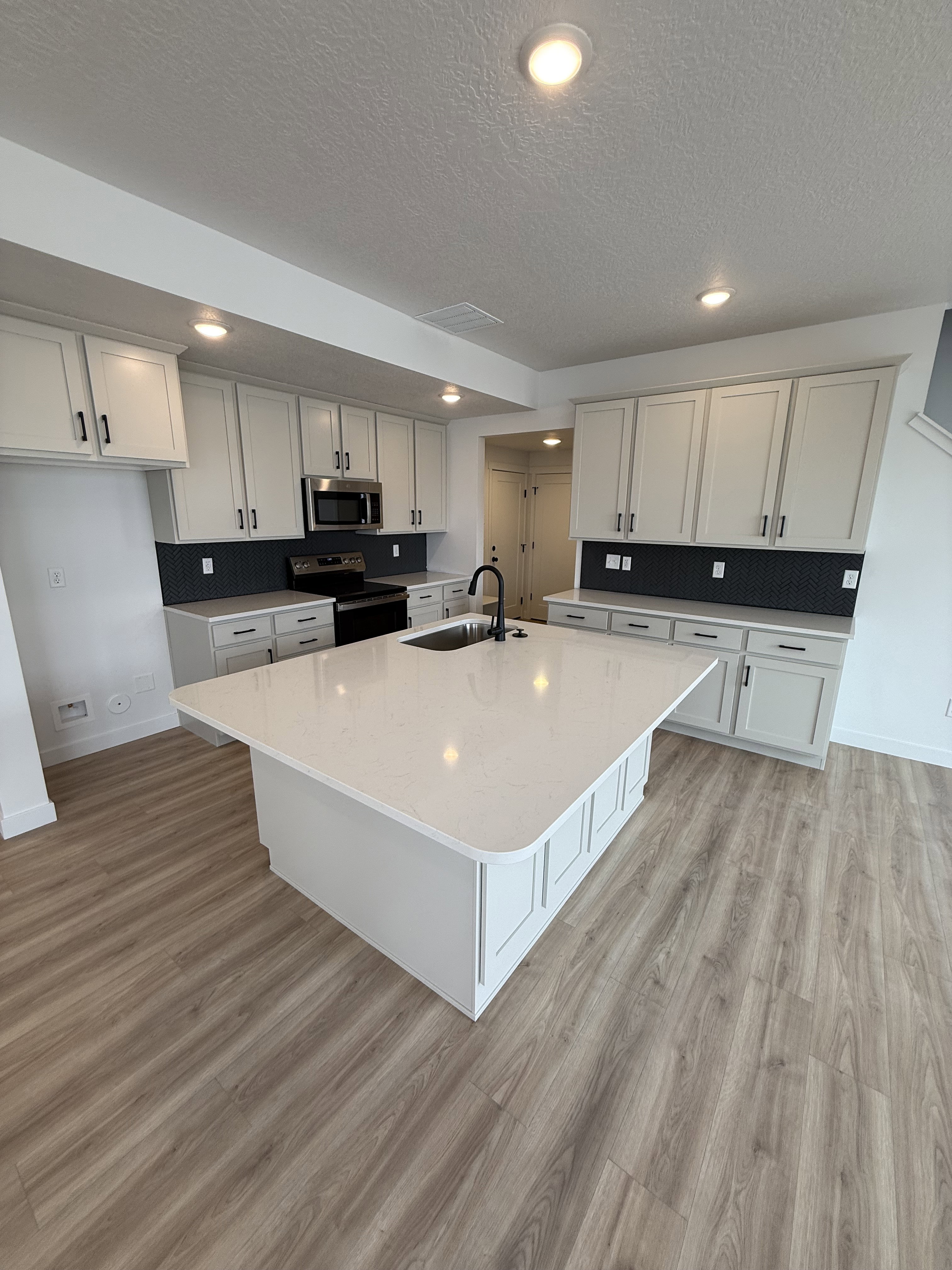 Kitchen featuring an expansive center island with integrated sink, light-toned cabinetry with dark hardware, a dark subway tile backsplash, stainless steel appliances, and wood-finish flooring