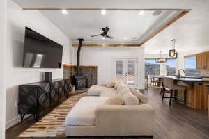 Living area featuring dark wood-type flooring, a ceiling fan, a wood stove, a raised ceiling, and french doors