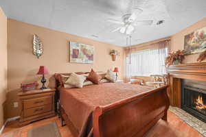 Bedroom featuring light wood finished floors, a textured ceiling, a fireplace with flush hearth, and a ceiling fan