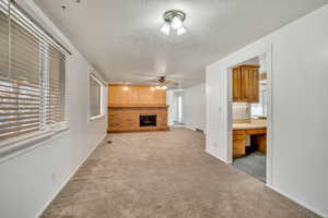 Unfurnished living room with a brick fireplace, a textured ceiling, built in desk, light carpet, and a ceiling fan