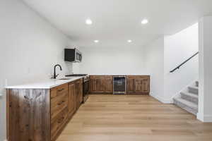 Indoor wet bar featuring beverage cooler, light wood-style flooring, stainless steel appliances, wood finish cabinetry, and light stone counters