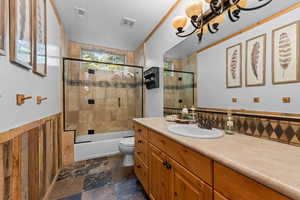Bathroom featuring vanity, shower / bath combination with glass door, wainscoting, and dark stone finish flooring