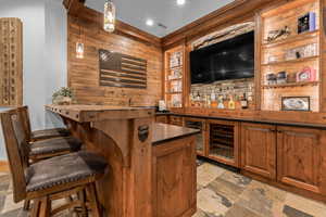 Indoor wet bar with wood finish cabinetry, wine cooler, hanging light fixtures, stone tile flooring, and wooden walls