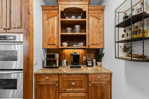 Bar area featuring double oven, open shelves, light stone counters, and wood finish cabinets