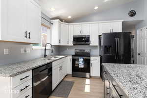 Two tone kitchen with light stone counters, black appliances, lofted ceiling, light wood-type flooring, and dual tone cabinets