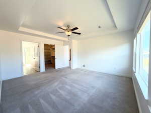 Unfurnished bedroom with a walk in closet, a tray ceiling, light colored carpet, and ceiling fan