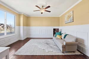 Living area with a decorative wall, ornamental molding, wainscoting, dark wood-style floors, and ceiling fan