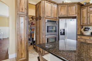 Kitchen featuring stainless steel appliances, dark stone countertops, wood finish cabinetry, arched walkways, and light tile patterned flooring