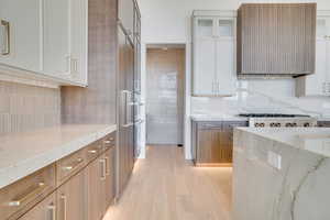 Two tone kitchen with dual tone cabinets, light wood finished floors, ventilation hood, light stone counters, and glass insert cabinets