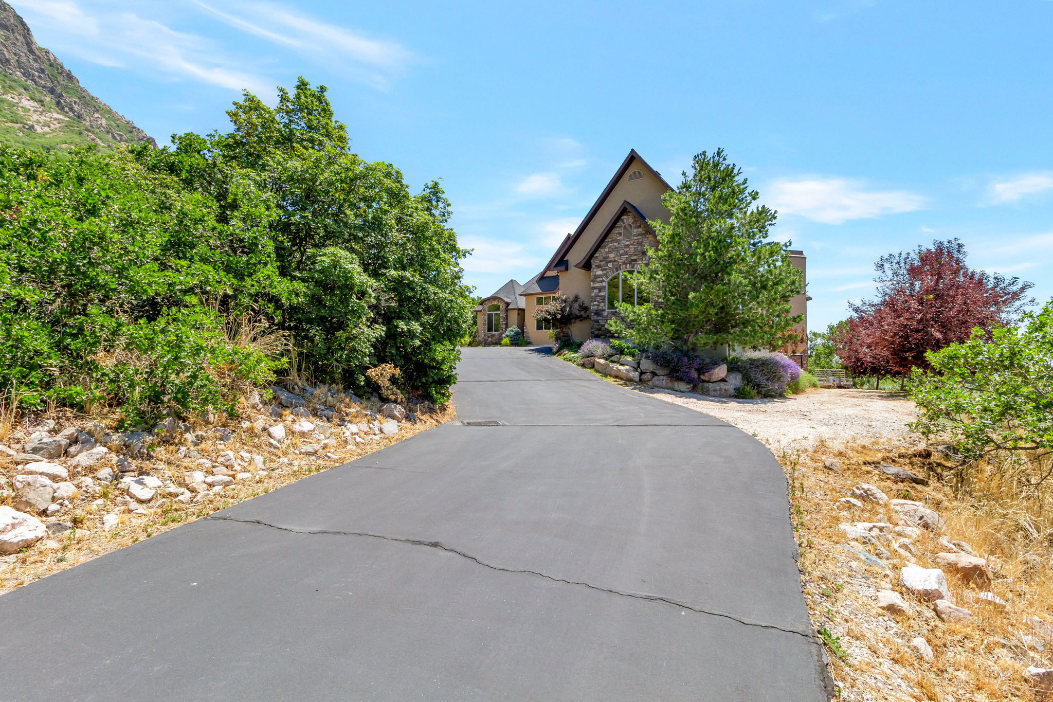 Curved asphalt driveway ascending to a stucco and stone-clad residence, featuring a peaked roofline and mature landscaping