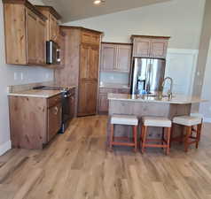 Kitchen with lofted ceiling, stainless steel appliances, a kitchen bar, an island with sink, and light stone countertops